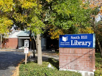 Blue South Hill Library sign on a brick base beside a tree-lined driveway with fall leaves