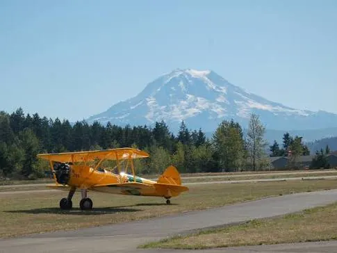 Yellow vintage biplane parked on the grass beside a taxiway at Thun Field with Mount Rainier behind the trees